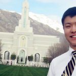 An LDS missionary of Asian decent is seen posing in front of an LDS temple with mountains in the background