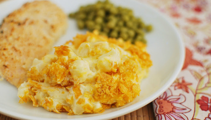A plate with funeral potatoes, peas and grilled chicken is shown with the potatoes in focus and the rest blurred in the background.