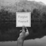 Hand holding card in nature with "Forgive Yourself" text on it.